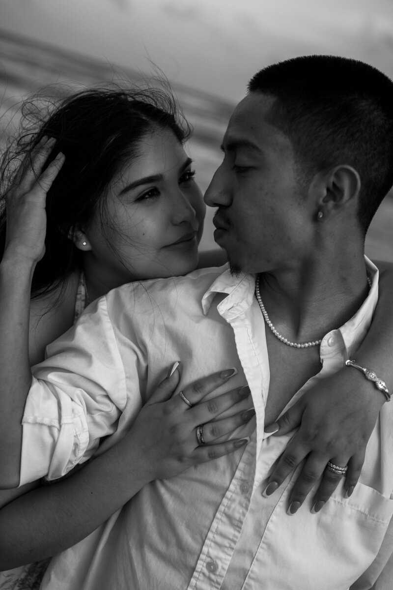 A romantic black-and-white photo of a couple on a beach. They embrace closely, gazing into each other's eyes, conveying love and intimacy.