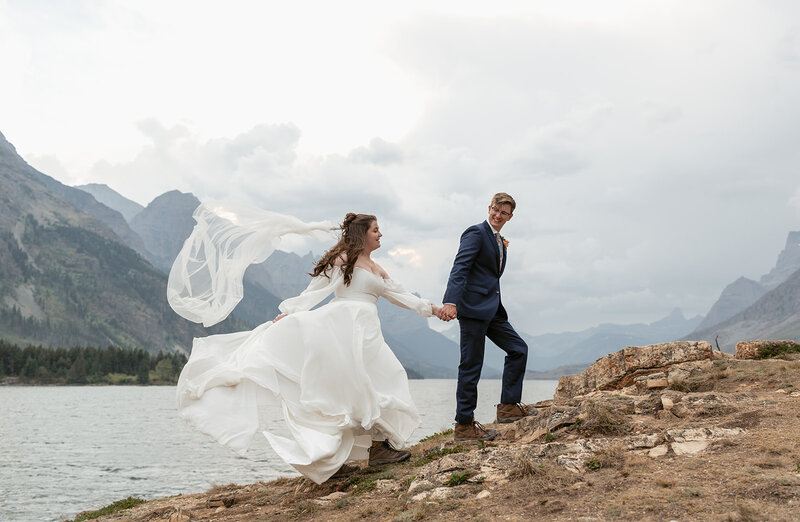 A joyful bride and groom run along the rocky shoreline of Glacier National Park, her flowing white dress and veil catching the mountain wind as they laugh together beside the lake, photographed by Sydney Breann Photography.