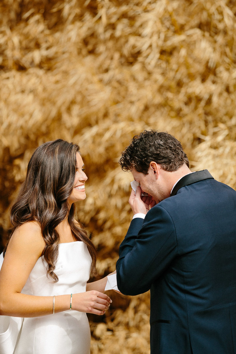 bride in strapless wedding dress smiling at groom in blue suit wiping away tears surrounded  by trees with orange leaves
