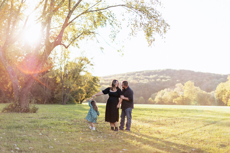 Family standing in the grass with parents looking at one another while daughter twirls and dances holding mom's hand at sunset. 
