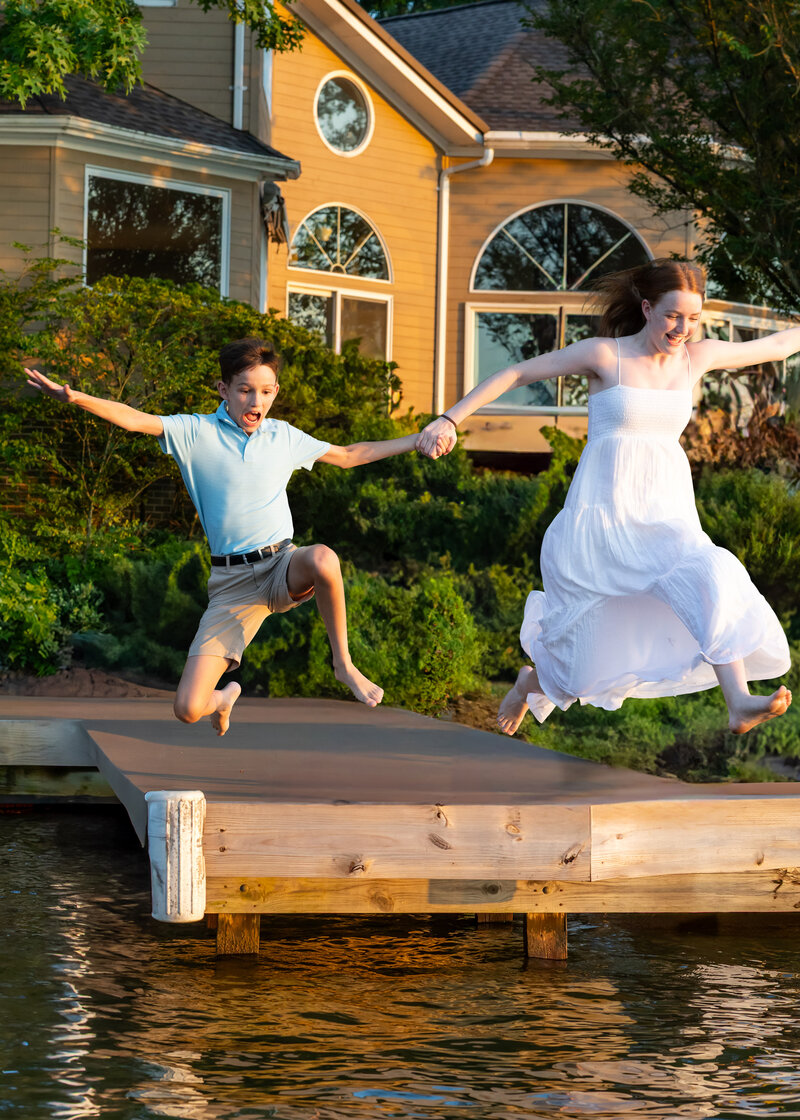 Brother and sister holding hands as they leap off a wooden dock into the water at sunset – captured by Capture Joy Photography, Northern Virginia family photographer