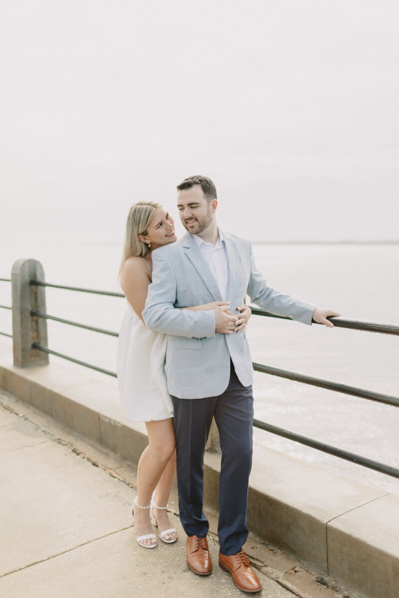 Bride in short white dress hugs groom in navy slacks and a baby blue sport coat from behind smiling over his shoulder. They are standing next to the guardrails of the Charleston Battery walkway on the river in Charleston, South Carolina.