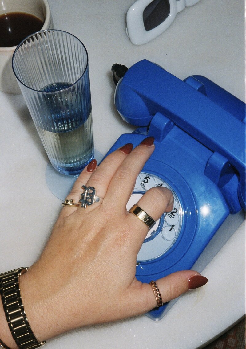 Hand with gold rings and red nails dialling an old fashioned blue phone with a glass of water beside the phone