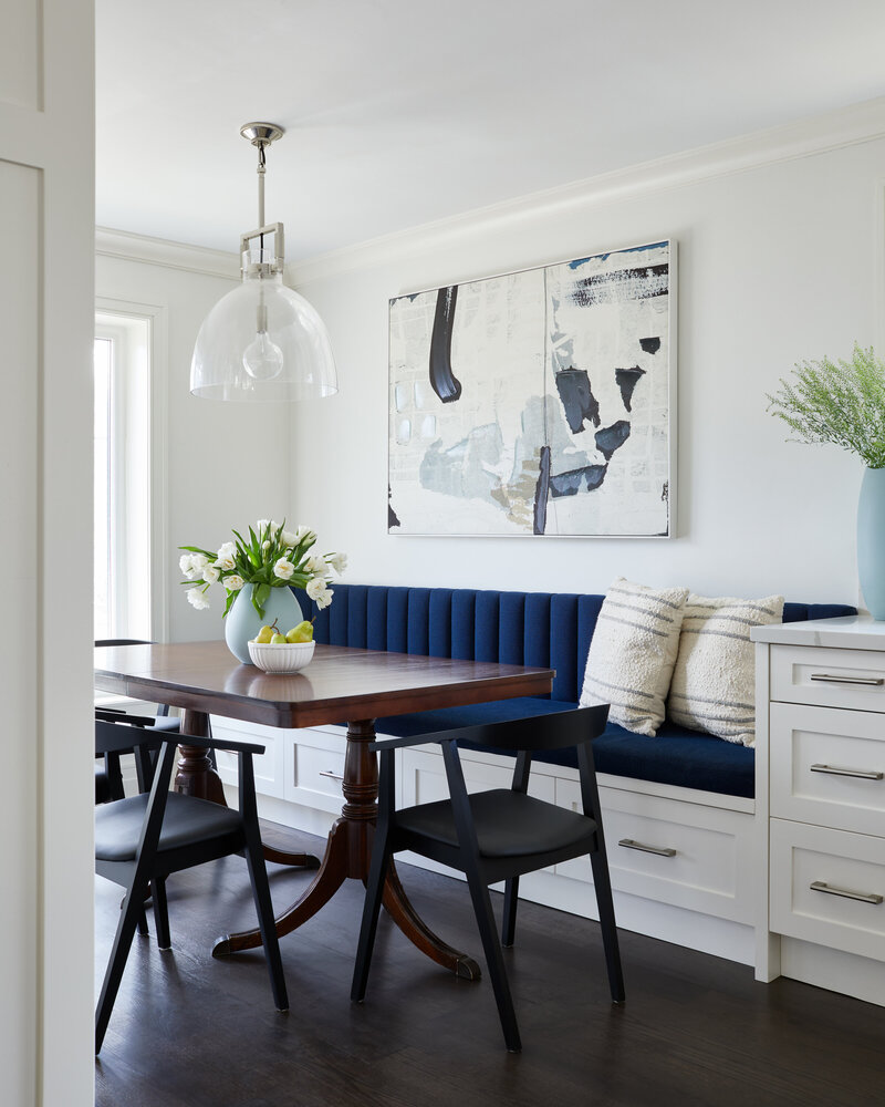 An elegant wood dining table in front of a custom banquette with blue channeled tufting in a brightly lit room with abstract wall art, potted plants, and a bowl of fruit