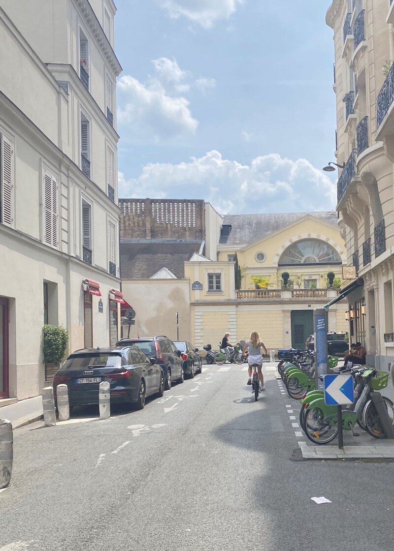 Jenn Cloutier's daughter cycling through Paris streets with historic architecture and bike share stations, representing self-leadership in action through intentional life experiences.