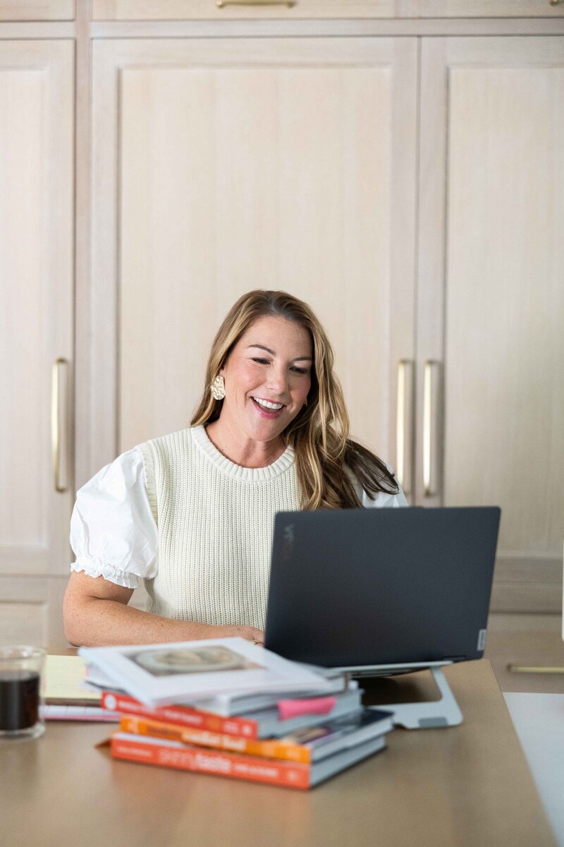 Woman with dark hair in cream shirt on laptop with cookbooks