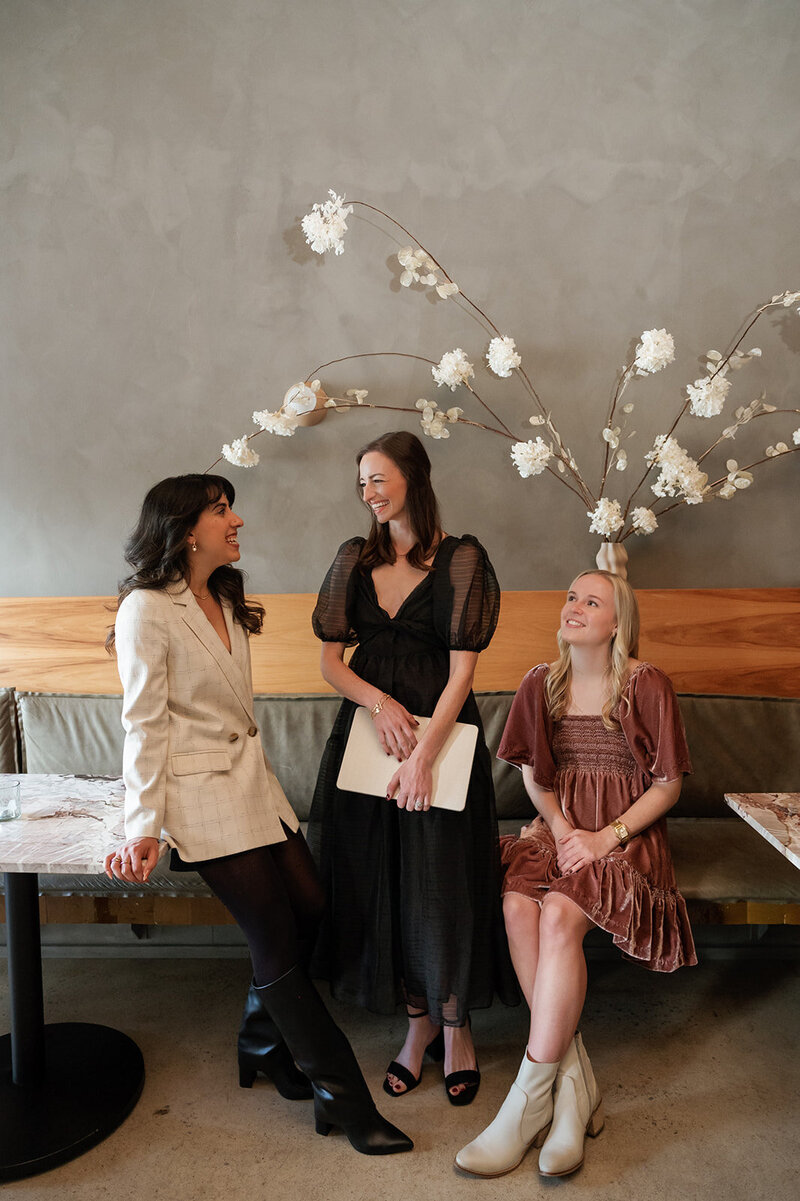 Three brand and web designers looking at each other posing in front of a wall with a large floral arrangement on a shelf