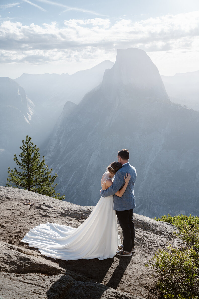 A couple in wedding clothes embraces and looks out at Half Dome in Yosemite National Park