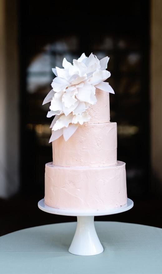 Atlanta wedding planner, Tyler Albritton's wedding cake from her wedding at the Dallas Arboretum & Botanical Gardens. The cake is three tiers, with pastel pink frosting, and white florals cascading down the first two tiers on the left side of the cake