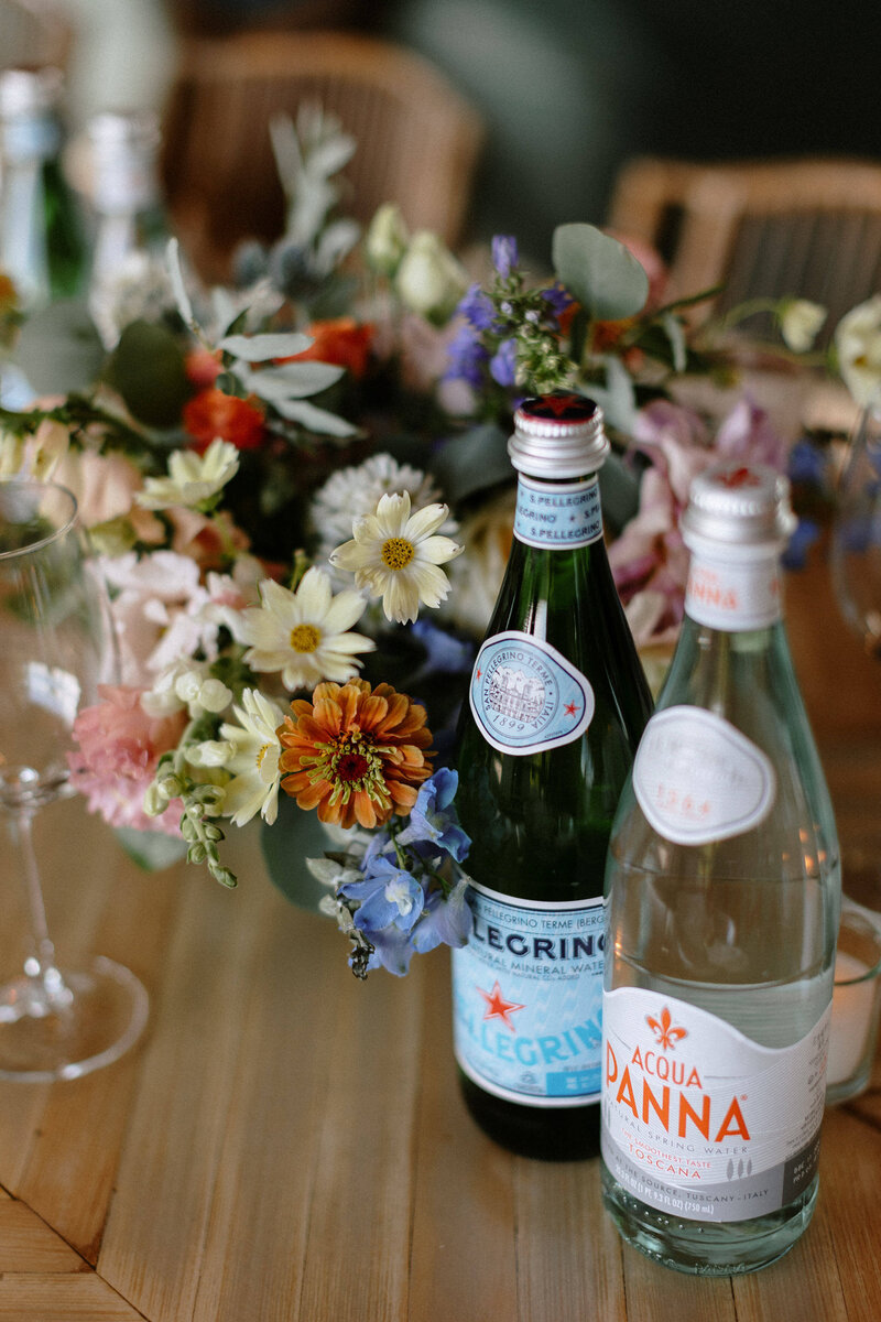 Two bottles of sparkling water, San Pellegrino and Acqua Panna, are surrounded by a vibrant floral arrangement on a wooden table.
