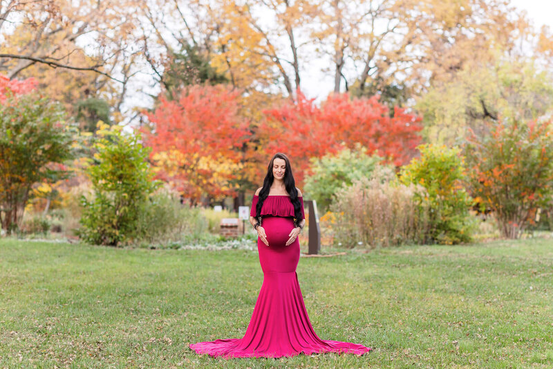 A pregnant woman in a red dress hugging her belly in front of a garden in Chantilly, VA.