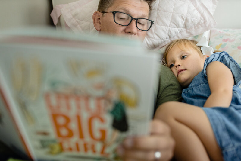 A little girl listens to her dad read her a book while laying next to one another.