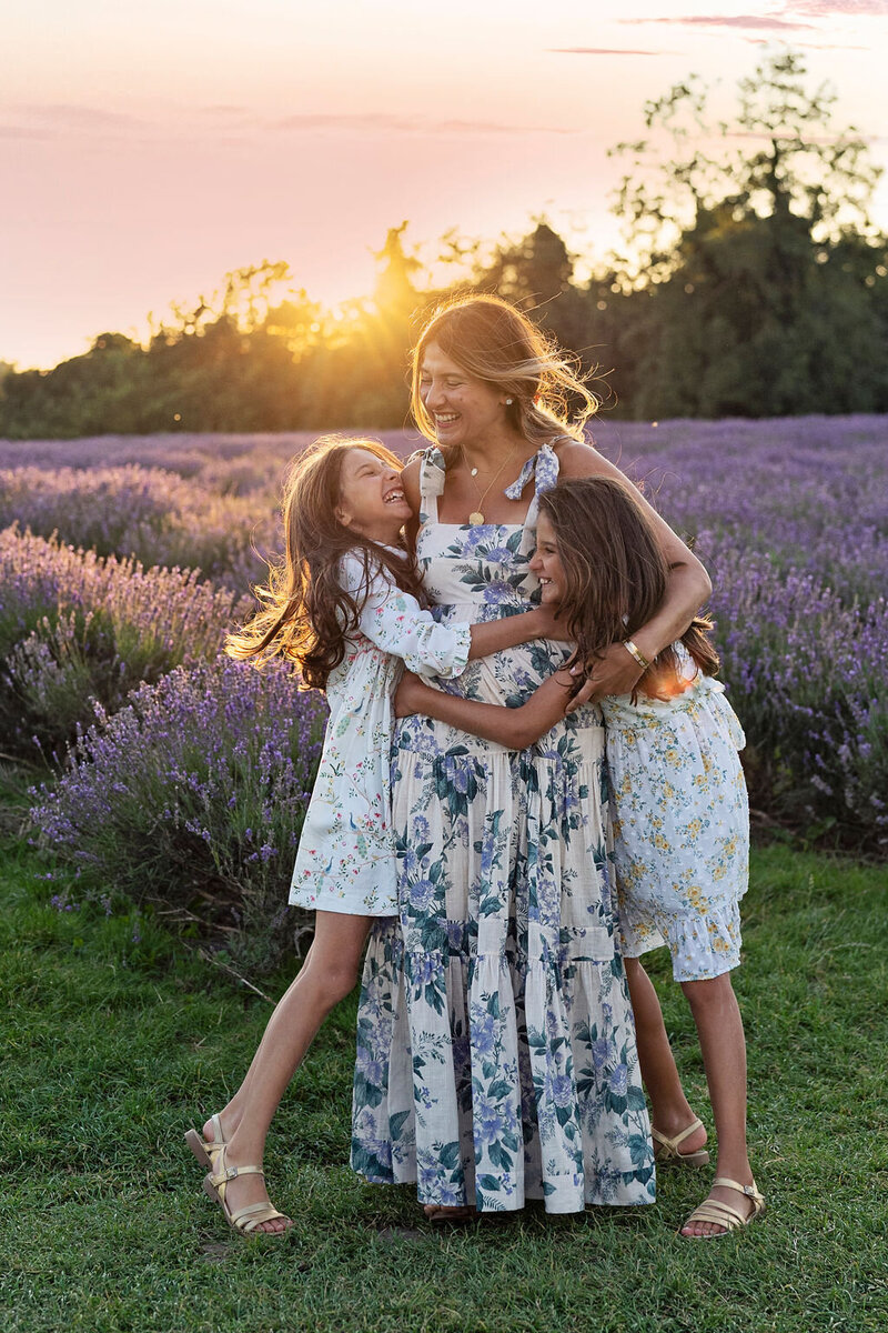 A mother is embraced by her two daughters in a lavender field as the sun sets in the background.