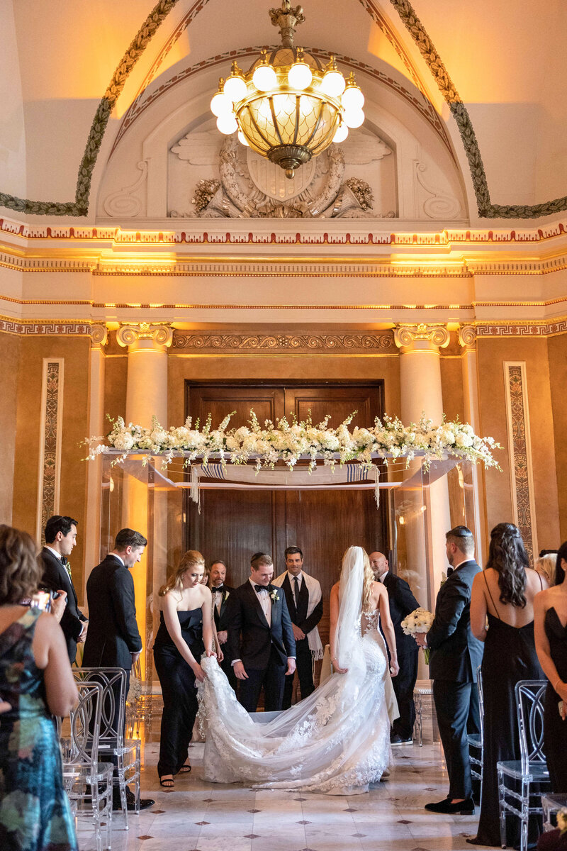 A wedding ceremony in an elegant, ornately decorated venue. The bride in a white gown and the groom in a black suit stand under a floral arch. Guests in formal attire surround them, with a chandelier overhead and marble columns in the background.