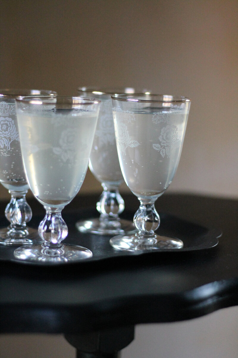 Elderflower wedding welcome cocktails in vintage glasses sit on a black tray resting on a table