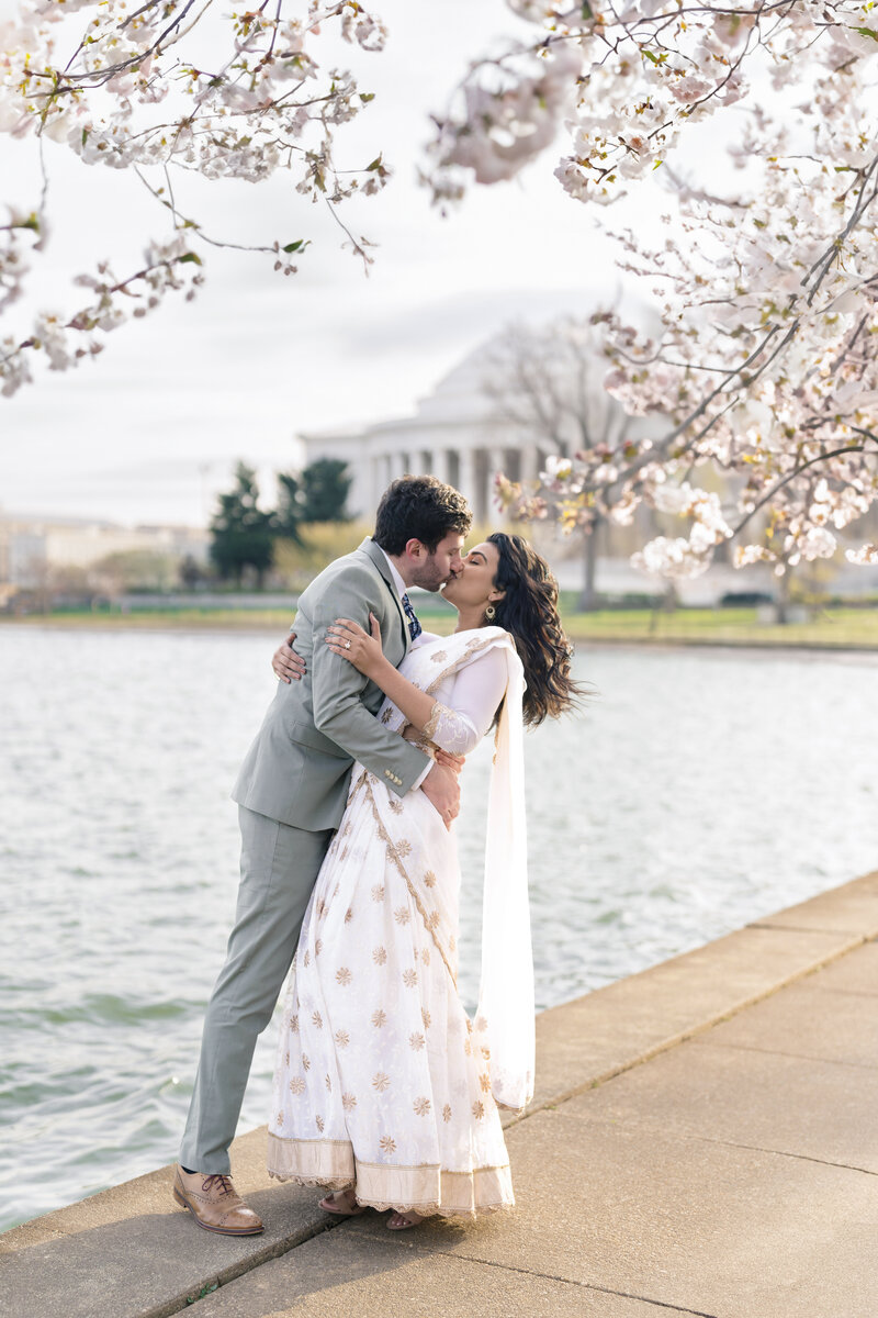 A couple kisses under cherry blossom trees by a waterfront. The woman wears a white and gold traditional dress, and the man wears a light gray suit. They are surrounded by blooming flowers and the water shines in the background.