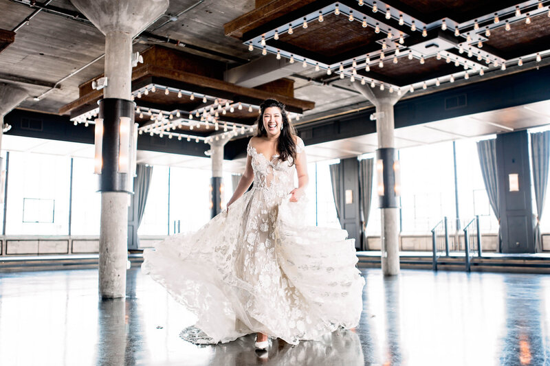 A smiling bride runs towards the camera, her dress flowing, in an industrial loft space.