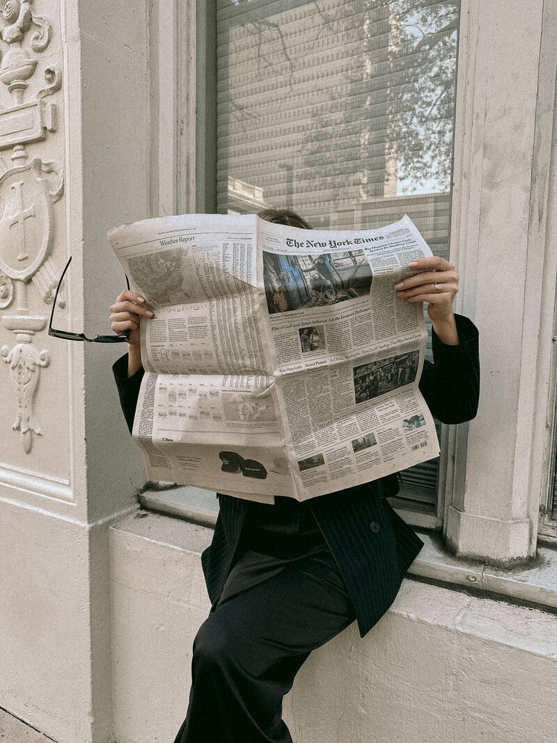 Woman reading The New York Times newspaper while sitting on stone steps, wearing dark suit.