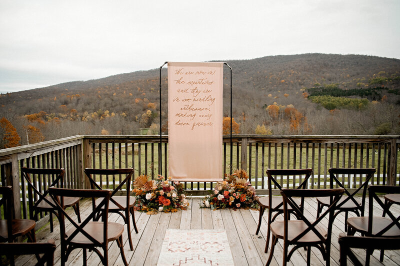 A large blush pink banner with flowers around the base stands on a deck overlooking the Catskill mountains. It reads "We are now in the mountains and they are in us, kindling enthusiasm, making every nerve quiver."