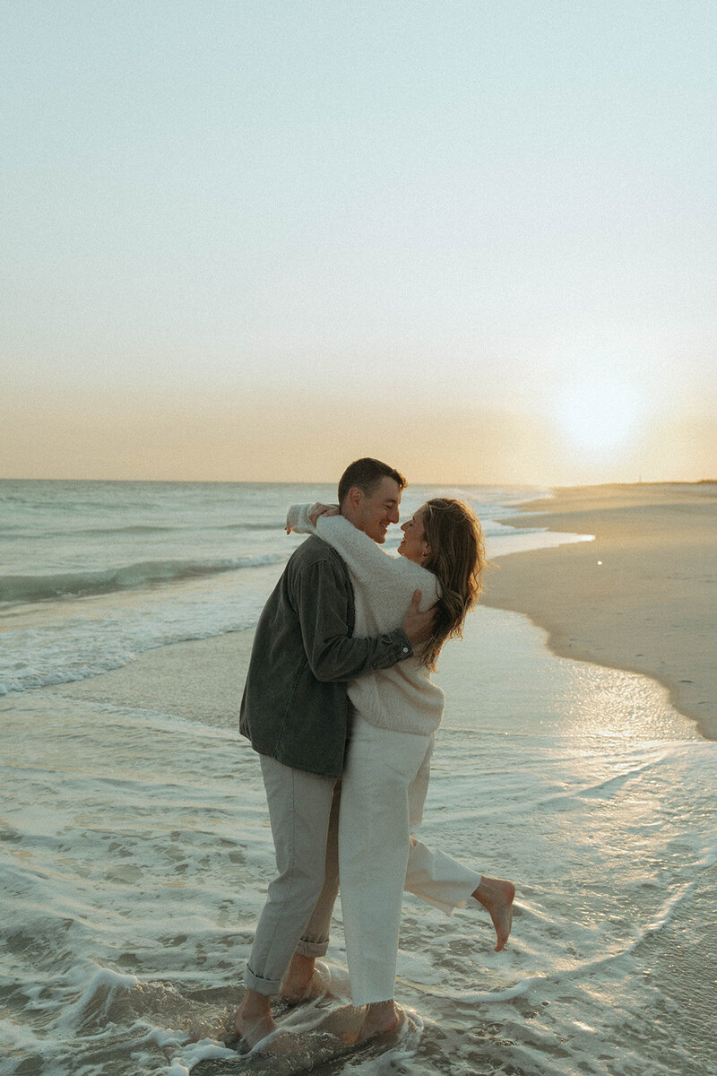 couple with feet in the waves and arms around each other during engagement photos at the beach, captured by Elsie Goodman, an NYC engagement and couples photographer