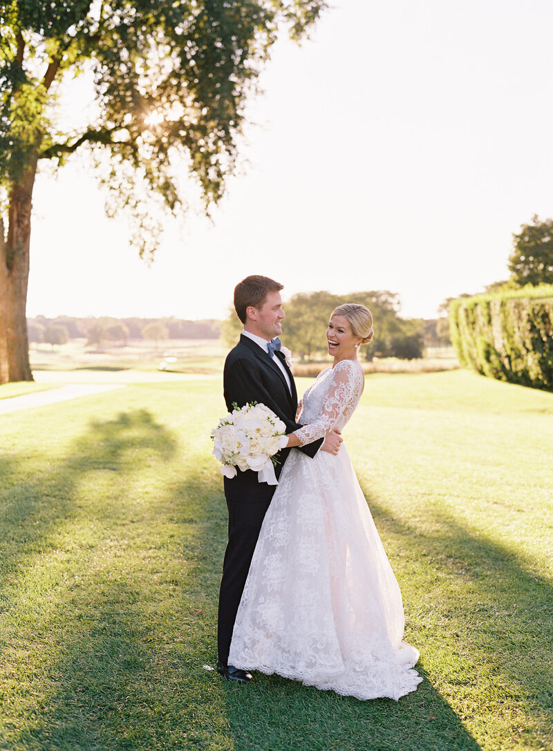 Newlyweds walk hand-in-hand across a sunlit, verdant meadow, with expansive trees in the background under a clear sky, capturing a serene and romantic moment.