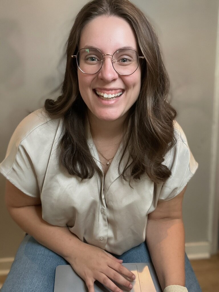 Smiling woman with glasses and brown hair, wearing a light blouse and jeans, sitting and looking at the camera. Molli Gorrell, Virtual Assistant and Web Designer.