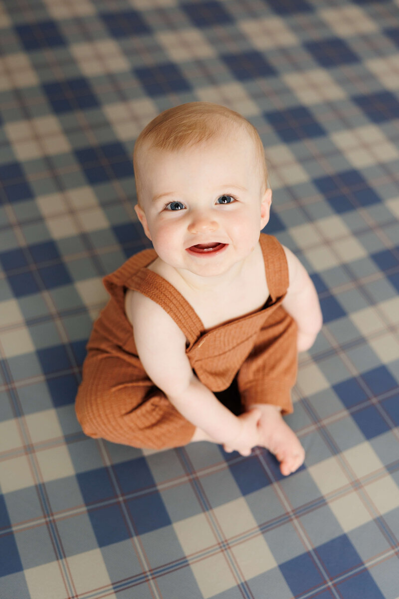 little boy sitting on blue plaid backdrop smiling up at the camera for first birthday cake smash portraits