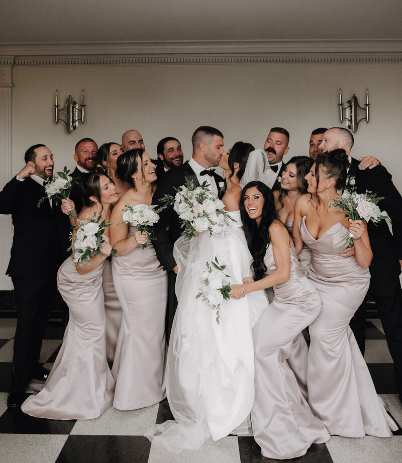 bride and groom kiss surrounded by their wedding party cheering them on at their new haven lawn club wedding photo by cait fletcher photography
