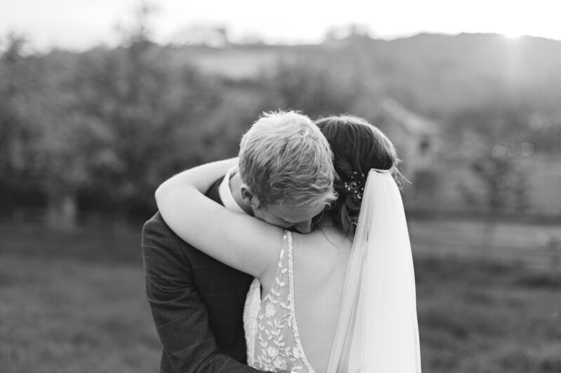 A black and white photo of a bride and groom embracing closely outdoors. The grooms head rests on the brides shoulder, and her veil cascades down her back. Trees and soft light create a serene backdrop.