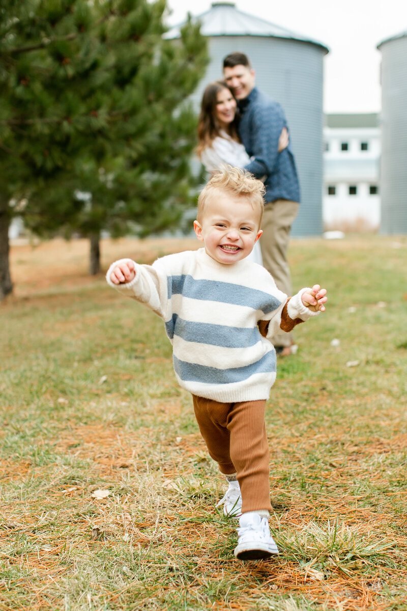 A little boy running toward the camera. His parents are hugging in the background.