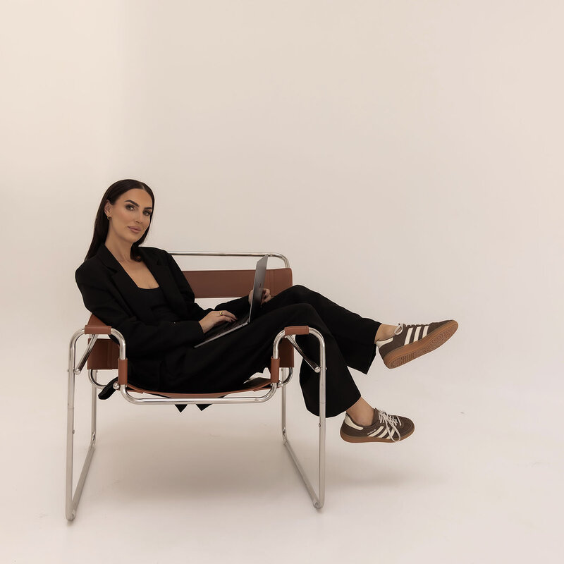 Tayla Bayne, founder of Aurum Design Studio, seated in a modern chair with a laptop and coffee, representing the brand’s story and creative roots.