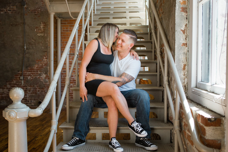 Happy husband and wife whisper to each other while sitting on industrial staircase in front of brick wall while dressed casually for photoshoot at Sky 9 Studio