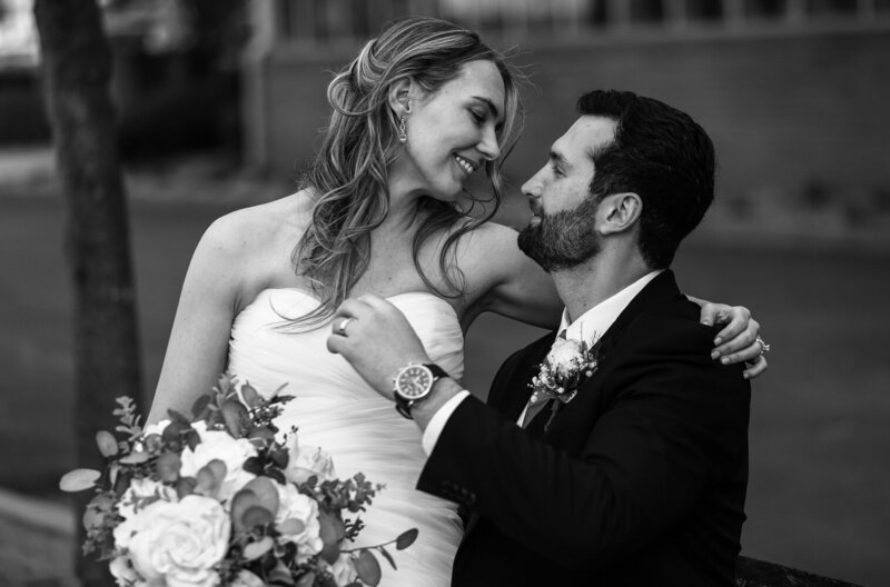 Bride sits on groom's lap with her arm around his shoulder  on a bench on walkway on Erie's Dobbins Landing