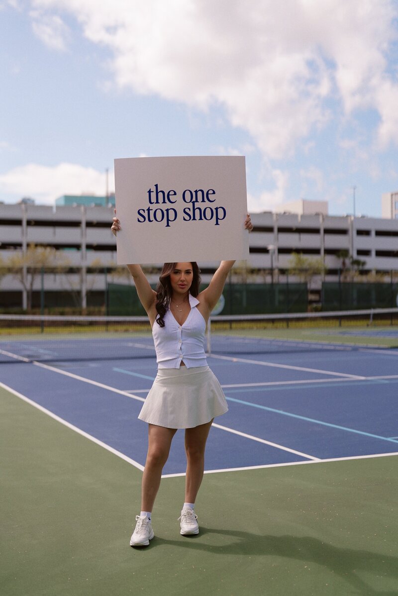 Woman on tennis court holding sign saying the one stop shop