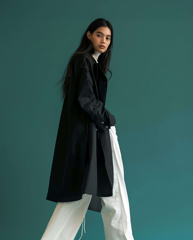 young woman in a long black coat and white pants walking to the right looking directly at us