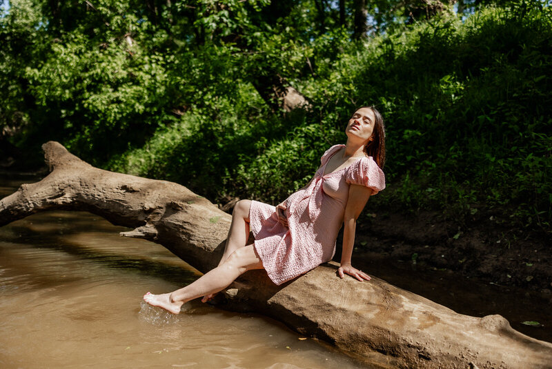 Expecting mother wearing a pink dress sitting on a fallen log in a river while holding her stomach and facing the sun with her eyes closed. 