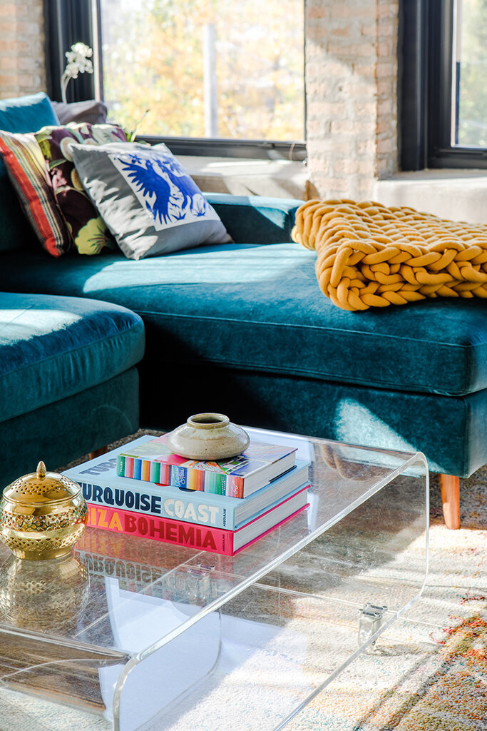 A close-up of a modern living room area. A clear acrylic coffee table holds a stack of colorful books titled "Turquoise Coast," "La Bohemia," and another with a blue and white spine. A small, textured ceramic pot sits on top of the books, and a decorative gold container rests beside them. A plush teal velvet sectional sofa with patterned pillows and a chunky mustard yellow knit blanket is partially visible in the background. Sunlight streams in from a window, casting shadows on the patterned rug underneath the coffee table.