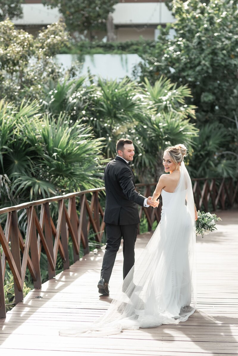 Bride in a fitted gown and groom in a black suit walking across a wooden bridge surrounded by tropical greenery, smiling back at the camera.