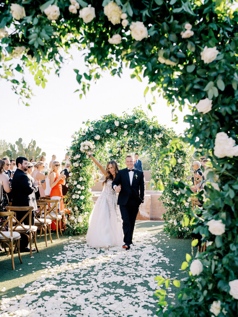 The bride and groom make a joyful exit under a breathtaking floral arch at their luxury Arizona Biltmore wedding. Surrounded by lush greenery and elegant white roses, this outdoor ceremony captures the beauty of a timeless celebration.