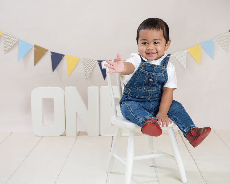 A baby boy in denim shorts and suspenders, with a navy polka dot bowtie, enjoying his cake smash session with frosting on his face and blue bunting in the background.