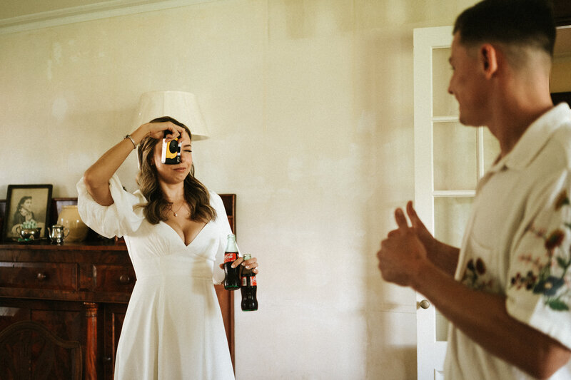 young bride taking photo of groom using 32mm film disposable camera, holding coke bottles