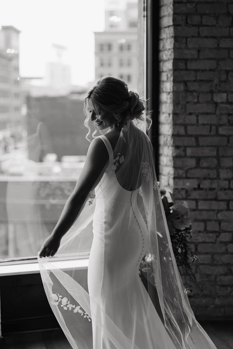 Elegant bride smiles down at her lacy veil while standing in front of window overlooking downtown Minneapolis