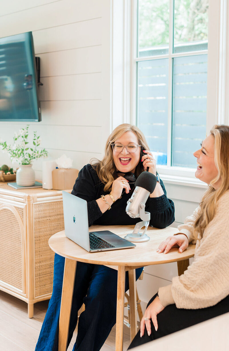 cory and melissa on a white background. melissa holding a laptop and corry holding a micrphone out to pretend to interview her