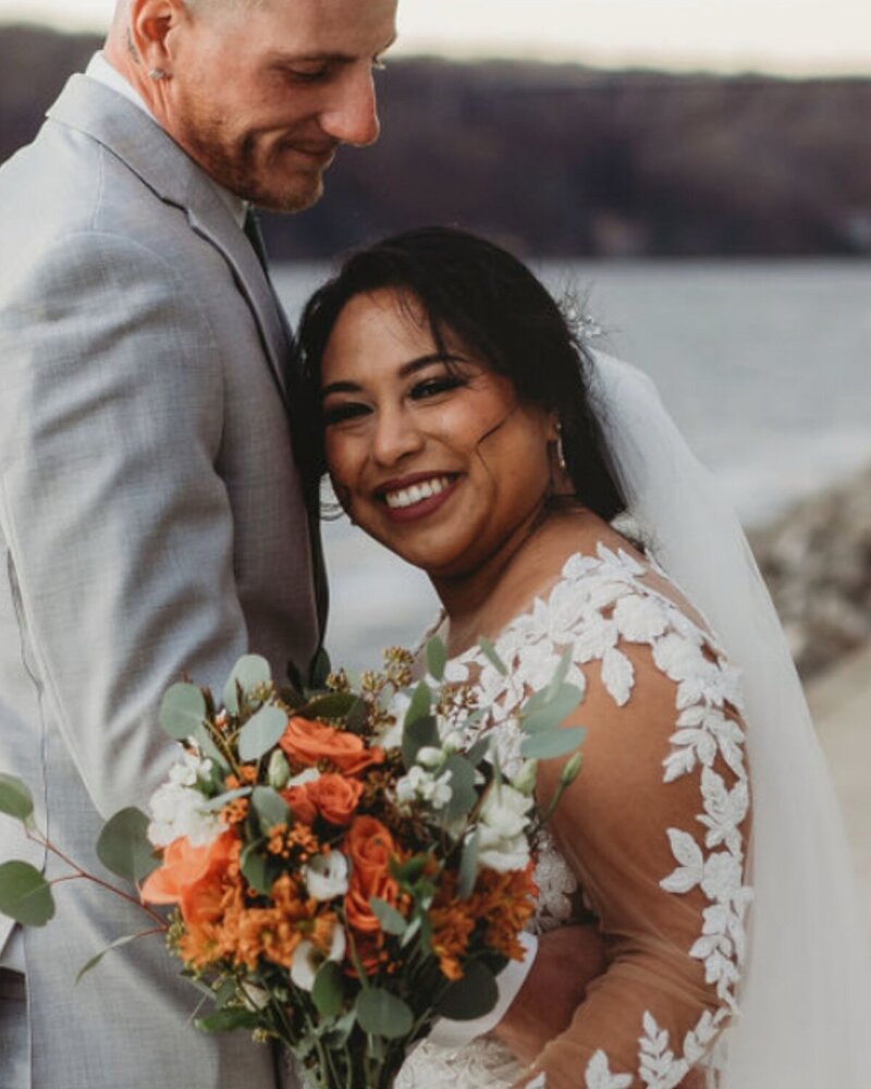 Outdoor photo of bride and groom by the water; the bride beams with bold lashes and timeless makeup—flawless wedding glam by Artistry by Jessica.