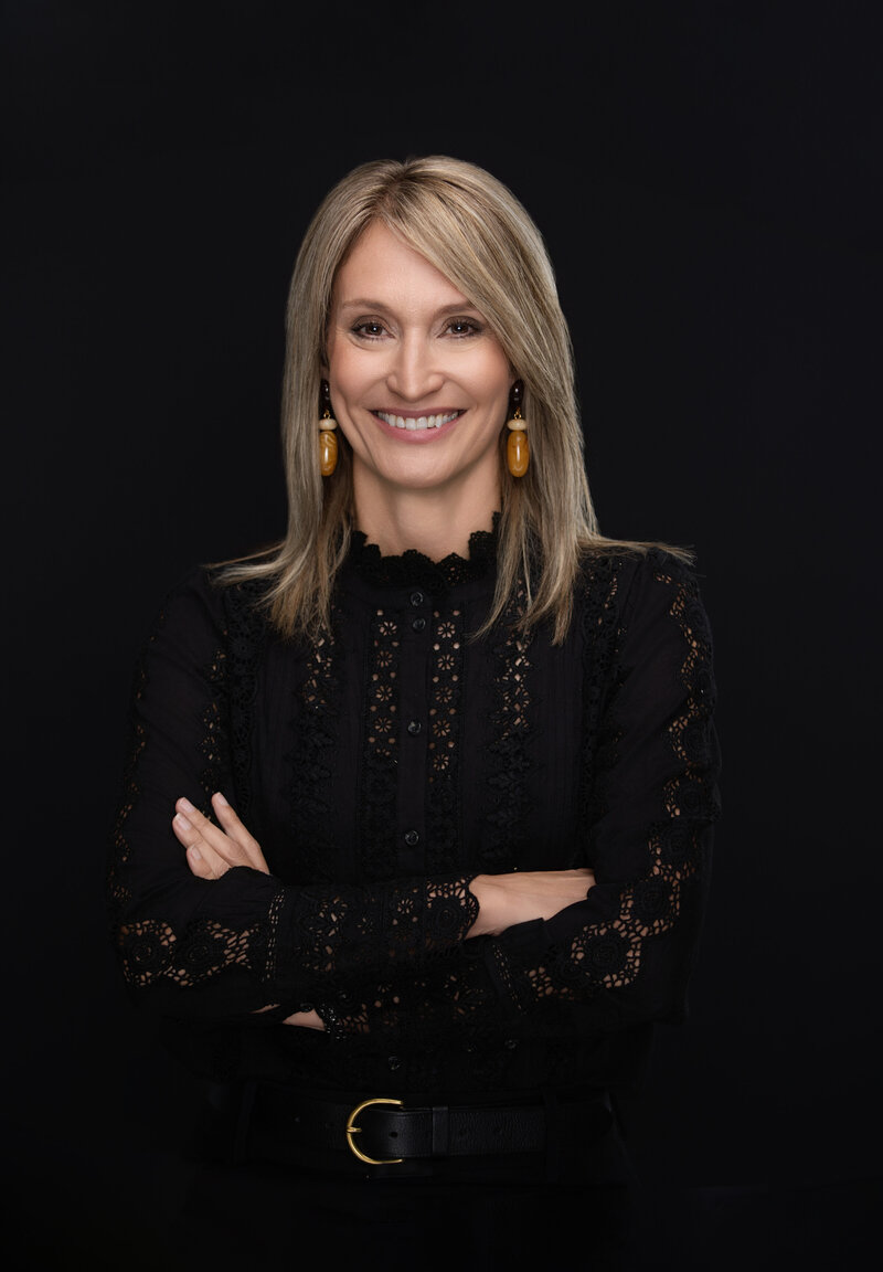 Professional headshot of a confident middle-aged woman with straight, shoulder-length blonde hair, photographed at Janel Lee Photography studio in downtown Cincinnati, Ohio. She is wearing a stylish black lace blouse with a high neckline and intricate embroidered detailing, complemented by bold amber drop earrings that add a modern, creative flair. Her arms are crossed in a relaxed, self-assured pose, and she’s smiling warmly with a sense of approachability and quiet strength. Captured against a seamless black backdrop, the image uses clean studio lighting to create subtle depth and dimension, ideal for corporate professionals or entrepreneurs seeking polished and personality-driven branding photos in Cincinnati. Perfect example of modern personal branding photography for women over 40.