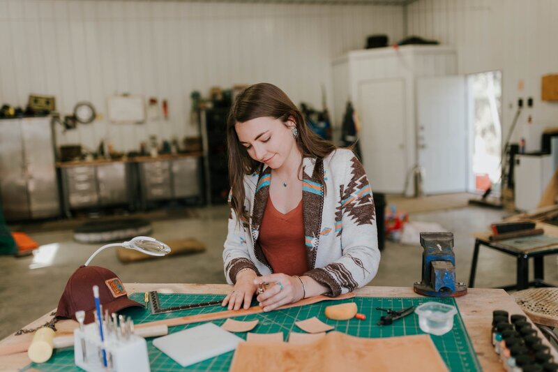 Woman in brown hair and wearing a  tribal print cardigan works at her desk creating handmade leather items during a branding photography session