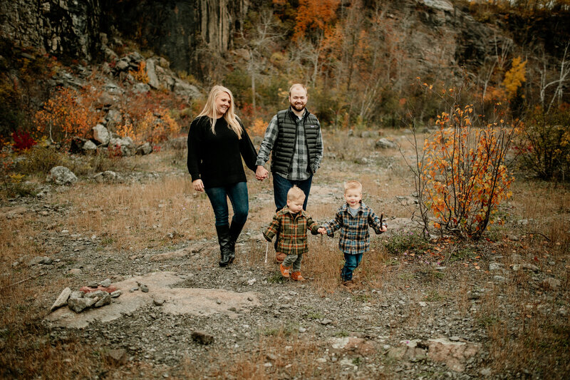 Two toddlers dressed in flannels run along hand-in-hand during Fall family photos while their parents trail behind.