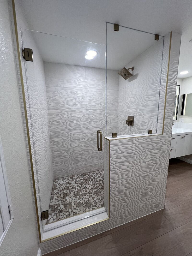 A fresh shower remodel with large white tiles that look like a wood grain texture and pebble looking tiles on the floor. New glass enclosure surrounds it.
