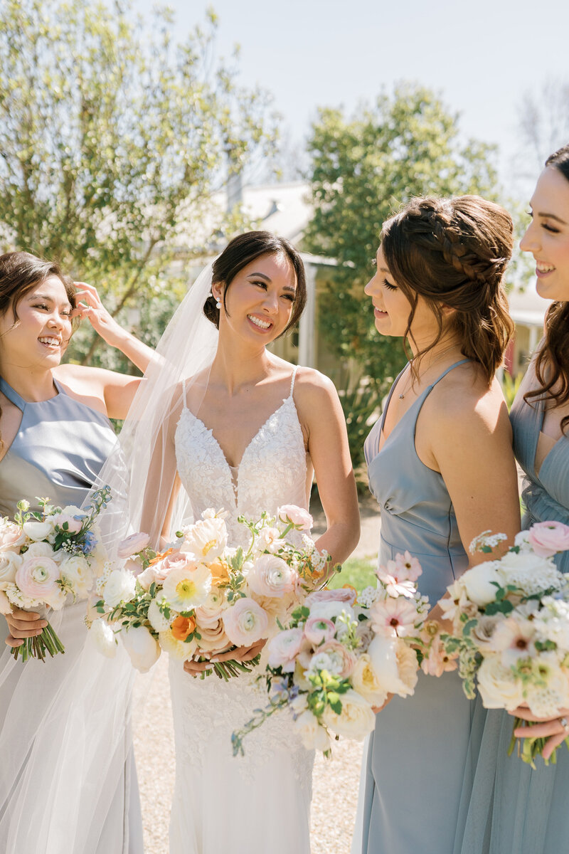 Bride smiling with her bridesmaids outside, all of them are holding light and lush flower arrangements