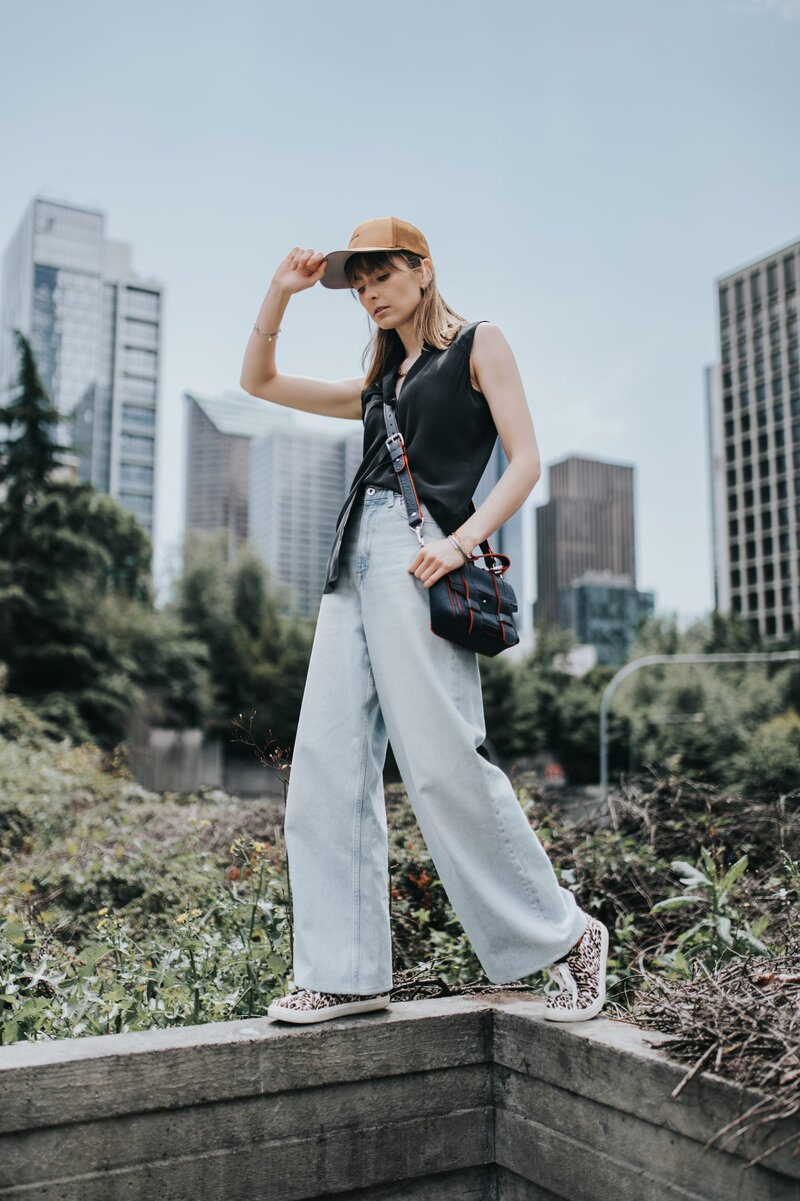 Woman in jeans and a cap stands on cement wall outdoors and poses casually while wearing a small, black purse.